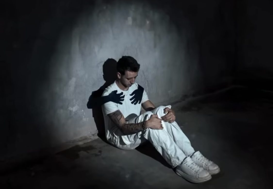 Man sitting alone in dark room with shadow hands on his chest, symbolising rumination, people-pleasing and men’s mental health struggles in Sydney