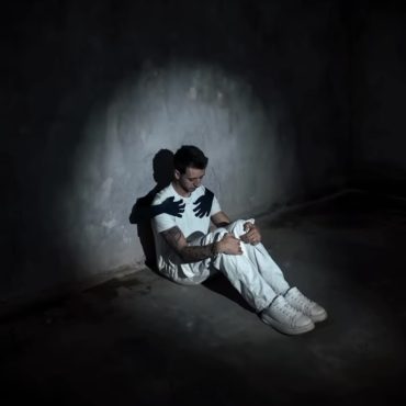 Man sitting alone in dark room with shadow hands on his chest, symbolising rumination, people-pleasing and men’s mental health struggles in Sydney