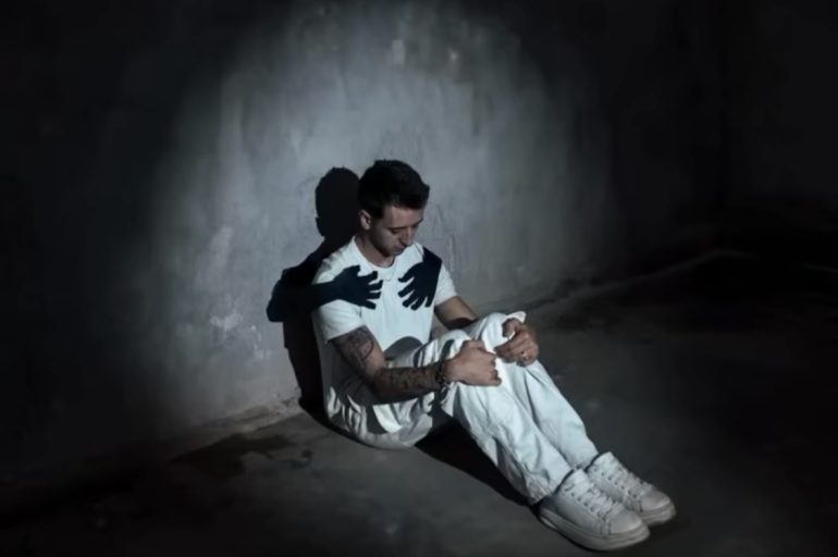 Man sitting alone in dark room with shadow hands on his chest, symbolising rumination, people-pleasing and men’s mental health struggles in Sydney